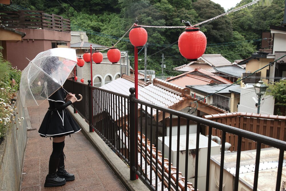 赤い提灯の並ぶ雨の温泉街を、ビニール傘をさして歩く人物